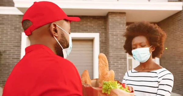 의료용 마스크를 쓴 아프리카계 미국인 남성 택배 원이 집에서 여성 고객에게 음식 가방을 전달하고 야외에서 음식을 나누어 주고 있습니다. 물건 꾸러미를 여자 손님에게 날라 주는 남자. 코로나 봉쇄. — 스톡 사진