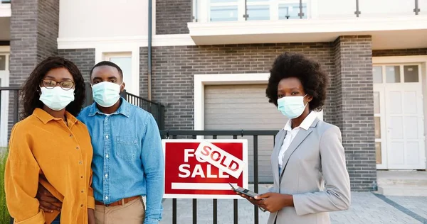 Afroamericano joven feliz pareja casada en máscaras médicas de pie al aire libre en la nueva casa con agente de bienes raíces femenino sonriendo a la cámara. Vendido a casa. Concepto pandémico. Compra de vivienda. —  Fotos de Stock