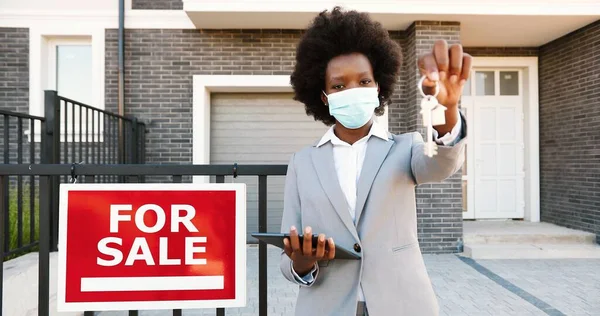 Retrato de joven afroamericana hermosa mujer agente de bienes raíces de pie al aire libre en casa con tabla En venta. Mujer en máscara médica que muestra y entrega las llaves de la vivienda en la periferia a la cámara —  Fotos de Stock