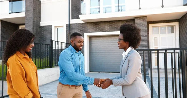 Imóveis femininos vendendo casa para casal feliz afro-americano e chaves de passagem. Ao ar livre. Homem e mulher recém-casados alegres sorrindo e comprando casa na periferia. Habitação em subúrbios. — Fotografia de Stock