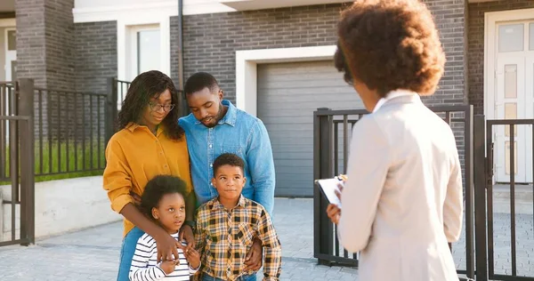 Giovane famiglia afroamericana con due bambini piccoli comprare casa in periferia e parlare con agente immobiliare femminile all'aperto. Coppia sposata con figli in cerca di abitazione nel quartiere periferico — Foto Stock