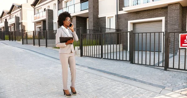 Afro-americano jovem agente imobiliário feminino em pé na rua no bairro suborb e falando no telefone móvel. Empresária com documentos à procura de casa e falando no celular. — Fotografia de Stock