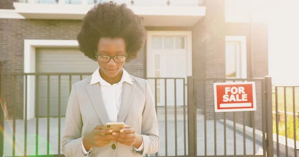 Ung afroamerikan vackra eleganta kvinnliga fastighetsmäklare i glasögon och affärsstil stående utomhus i utkanten på hus till salu och sms: a budskap på smartphone. Användning av mobiltelefon. — Stockfoto