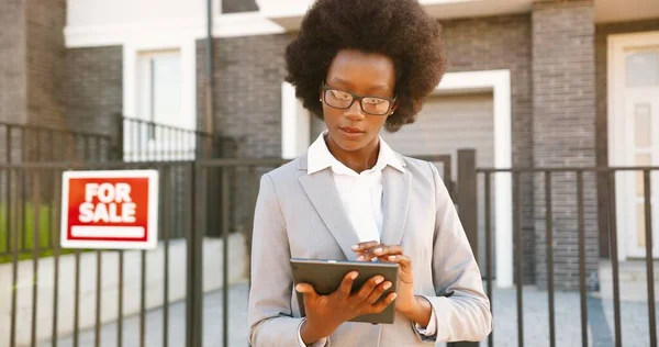 Ung afroamerikan vacker kvinnlig fastighetsmäklare i glasögon och affärsstil står utomhus i förorten på hus till salu och sms: a på surfplatta enhet. Använda gadget dator och knacka. — Stockfoto