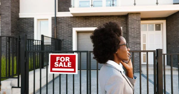 Afro-americano giovane elegante agente immobiliare femminile alla periferia di strada e parlando sul telefono cellulare. Donna d'affari in cerca di casa e che parla al cellulare. Abitazione in vendita. — Foto Stock