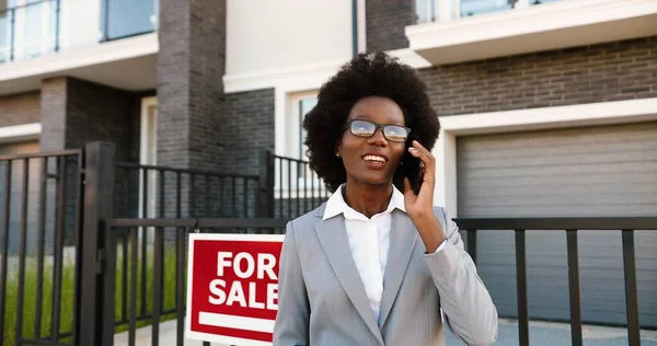 Afro-americano giovane elegante agente immobiliare femminile in occhiali nel quartiere periferico e parlando sul telefono cellulare. Donna d'affari in cerca di casa e che parla al cellulare. Abitazione in vendita. — Foto Stock