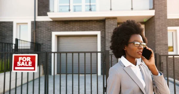 Afro-americano giovane elegante agente immobiliare femminile in occhiali nel quartiere periferico e parlando sul telefono cellulare. Donna d'affari in cerca di casa e che parla al cellulare. Abitazione in vendita. — Foto Stock