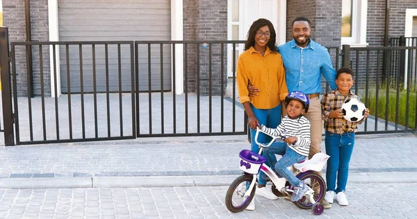 Ritratto di felice famiglia afroamericana con bambini piccoli in piedi in strada vicino casa al sobborgo e sorridente alla macchina fotografica. Madre allegra, padre, figlia e figlio carini. Genitori e figli. — Foto Stock