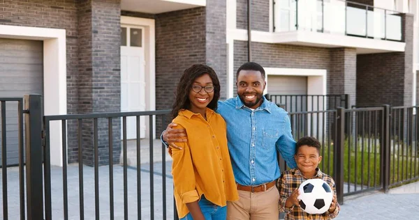 Porträtt av lycklig afroamerikansk familj med liten söt pojke som står på gatan nära huset vid förorten och ler mot kameran. Glada mor, far och son. Föräldrar och barn med fotboll. — Stockfoto