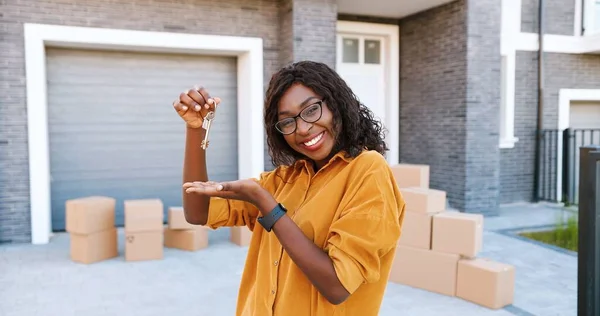 Πορτρέτο του χαρούμενα ευτυχισμένη Αφροαμερικανός γυναίκα σε γυαλιά χαμογελώντας στην κάμερα και δείχνοντας το κλειδί για την κάμερα, ενώ κινείται στο νέο σπίτι. Εξωτερικά. Γυναικεία κλειδιά επίδειξης. Κουτιά χαρτοκιβωτίων στο φόντο. — Φωτογραφία Αρχείου