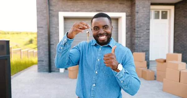 Portrait of cheerful happy African American man smiling to camera and showing key to camera while moving in new home. Outdoor. Male demonstrating keys. Carton boxes on background. Owner of real-estate — Stock Photo, Image