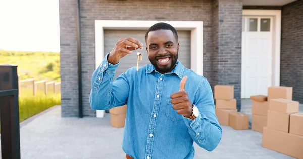 Ritratto di allegro uomo afroamericano felice che sorride alla macchina fotografica e mostra la chiave della fotocamera mentre si trasferisce in una nuova casa. All'aperto. Maschio che mostra le chiavi. Scatole di cartone su sfondo. Proprietario di beni immobili — Foto Stock