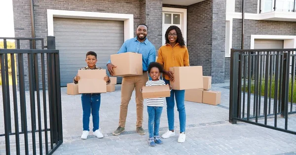 Retrato família afro-americana feliz com crianças pequenas em pé na casa nova no subúrbio e sorrindo com caixas de papelão nas mãos. — Fotografia de Stock