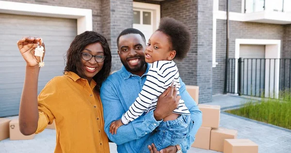 Portrait of happy African American family with small cute girl on hands standing at big house on suburb, smiling and showing key. Joyful mother, father with little pretty daughter. Moving in new home. — Stock Photo, Image