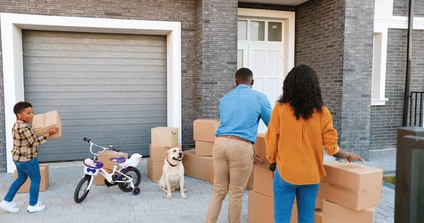 Afroamerikansk lycklig familj med små barn flyttar in i nytt hem i utkanten. Mor och far bär kartonger till gården och små barn hjälper till. Föräldrar med son, dotter och hund. — Stockfoto