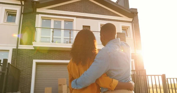 Arrière sur jeune couple afro-américain vient de se marier debout à la cour de la maison, étreignant et observant le deuxième étage. Vue arrière sur l'homme et la femme dans les câlins se déplaçant dans une nouvelle maison. — Photo