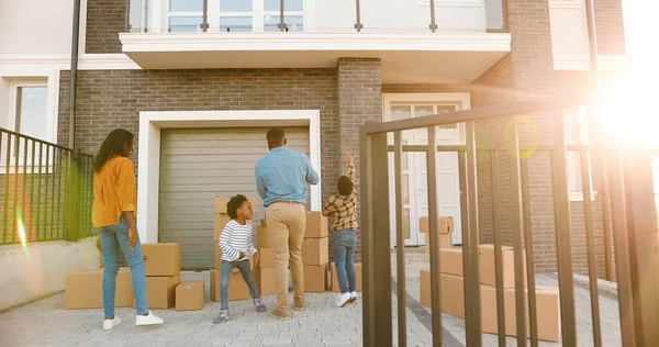 Afro-Amerikaanse gelukkige familie met kleine kinderen verhuizen in een nieuw huis. Vrolijke moeder en vader die lopen en kartonnen dozen naar huis dragen en kleine kinderen die helpen. Ouders met zoon, dochter. — Stockfoto