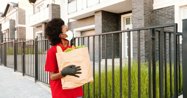 Afro-americana jovem mensageiro em máscara médica e luvas andando na rua e levando sacos com alimentos saudáveis. Mulher trabalhadora de parto passeando com pacote de legumes. Conceito de quarentena. — Fotografia de Stock