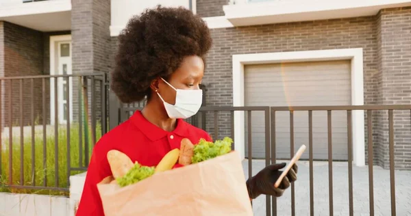 Donna afroamericana giovane consegna in maschera medica e guanti in piedi all'aperto con borsa piena di verdure e utilizzando smartphone per trovare la posizione. Il corriere donna sta intercettando il telefono. Fornire cibo. — Foto Stock