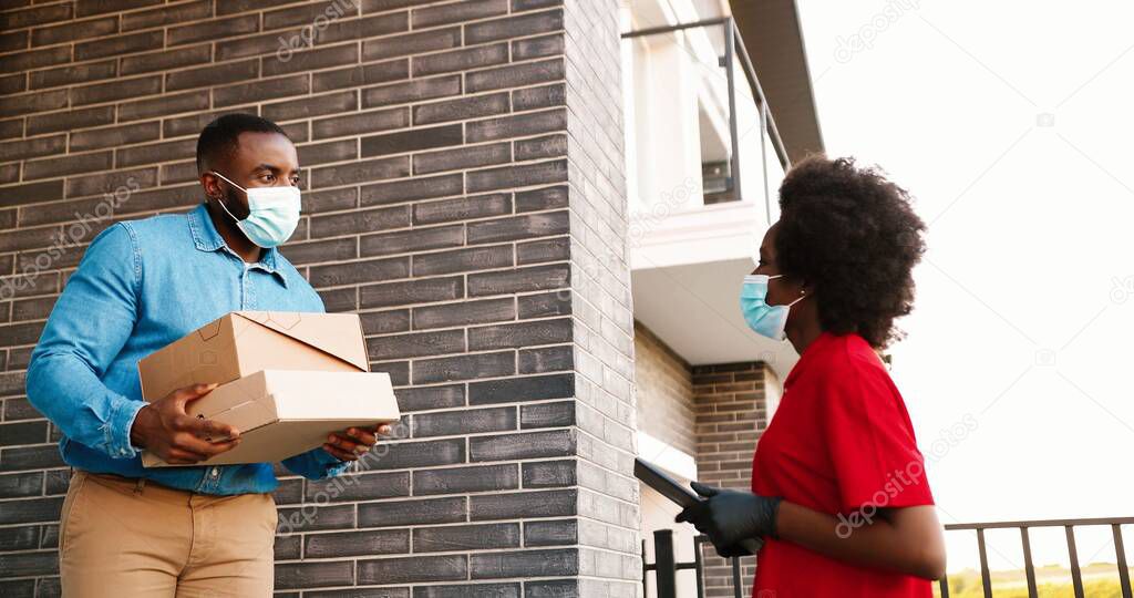 African American female courier in mask delivering carton parcels to male client at house and handing outside. Delivery woman bringing pizza boxes to man customer. Coronavirus concept.