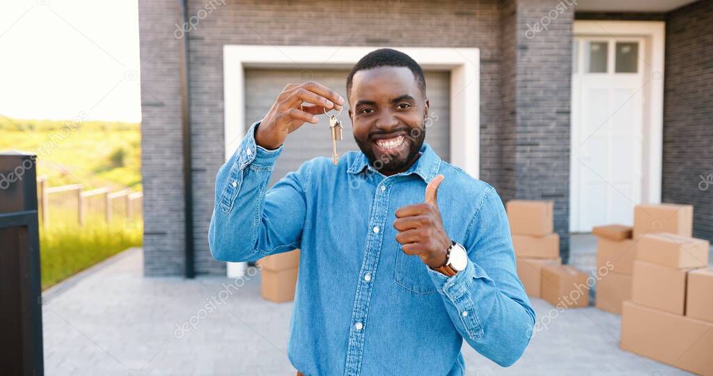 Portrait of cheerful happy African American man smiling to camera and showing key to camera while moving in new home. Outdoor. Male demonstrating keys. Carton boxes on background. Owner of real-estate