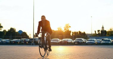 Beyaz tenli erkek bisikletçi gün batımında gülerdi. Bisiklet süren şık yakışıklı bir adam ve araba parkından geçiyor. Arabalar arka planda. Neşeli adam şehirde güzel bir yaz akşamında at sürüyor..