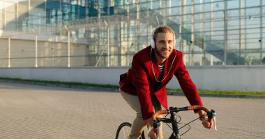 Beyaz, neşeli, şık, kırmızı ceketli ve kulaklıklı bisikletçi sokakta bisiklet sürüyor. Yakışıklı, neşeli, gülümseyen, bisiklet süren ve müzik dinleyen adam. Şehir manzarası