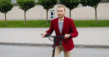 Açık havada elektrikli scooterlı ve kameraya gülümseyen kırmızı ceketli beyaz bir genç adam portresi. Şehir caddesindeki araçta şık görünümlü bir adam. Dolly vurdu. Yakınlaştırılıyor.