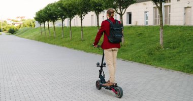 Şehir caddesinde elektrikli scooter kullanan sırt çantalı, beyaz, yakışıklı bir adamın arkası. Modern kasaba. Kırmızı ceketli, şık ve mutlu bir adamın araba sürüşü hakkında arka plan. Erkek sürücü.
