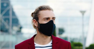 Dışarıdaki kameraya bakan maskeli beyaz, yakışıklı bir adamın portresi. Sokakta kulaklarında küpe olan şık, yakışıklı bir erkeğin yüzünü kapatın. Coronavirus salgını konsepti. Sosyal uzaklık