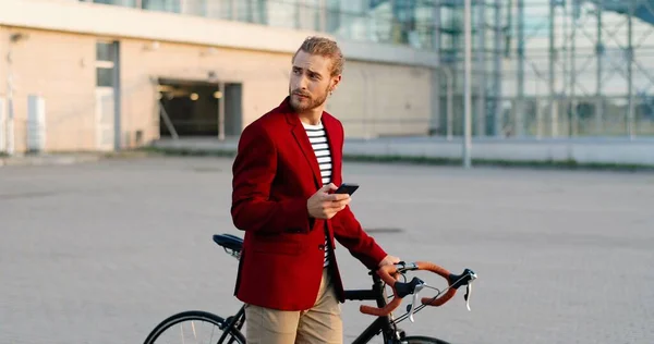 Caucasian male bicyclist in casual style and red jacket leading bike and tapping or scrolling on smartphone at street. Handsome young man walking with bicycle and texting message on mobile phone.