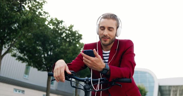 白种人英俊的年轻人 头戴耳机 靠在自行车或电动车上唱歌 英俊而时尚的男性在自行车上用智能手机听音乐 手机上的播放器 — 图库照片