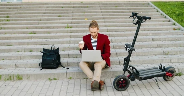 Kırmızı ceketli genç beyaz adam dışarıda merdivenlerde oturuyor, dizüstü bilgisayarda çalışıyor, içkisini yudumluyor. Kahve içen şık bir adam. Serbest çalışmakla meşgul. Elektrikli scooter. Bağımsız.