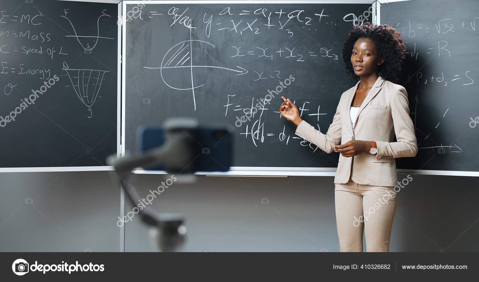 在学校，智能手机在网络摄像头上录制视频虚拟课程。在线学习。在黑板上教授数学或物理公式的非裔美国女讲师。封锁的概念。大流行病研究. —  图库照片©VAKSMANV101＃410326682