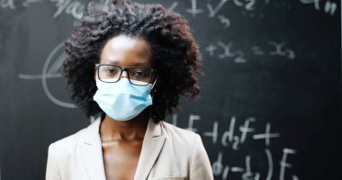 Sınıfta kameraya bakan gözlüklü ve tıbbi maskeli genç Afro-Amerikan bayan öğretmenin portresi. Arkaplanda formüller olan karatahta. Coronavirus konsepti. Pandemik okul.