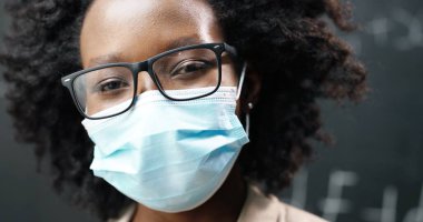 Gözlüklü ve tıbbi maske takmış kameraya bakan genç Afro-Amerikan bayan öğretmeni yakın çekim. Arkaplanda formüller olan karatahta. Coronavirus konsepti. Salgın hastalık eğitimi. Solunum koruması