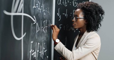 Okuldaki Afrikalı Amerikalı genç bayan öğretmen tahtaya formüller ve matematik kanunları yazıyor. Okul konsepti. Fizik kanunlarını açıklayan gözlüklü kadın matematik hocası. Eğitim.