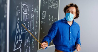 Tıp maskeli beyaz erkek öğretmen sınıfta ayakta duruyor ve fizik ya da geometri kanunlarını sınıfa anlatıyor. Salgın hastalık konsepti. Coronavirus sırasında okul. Eğitici matematik dersi.