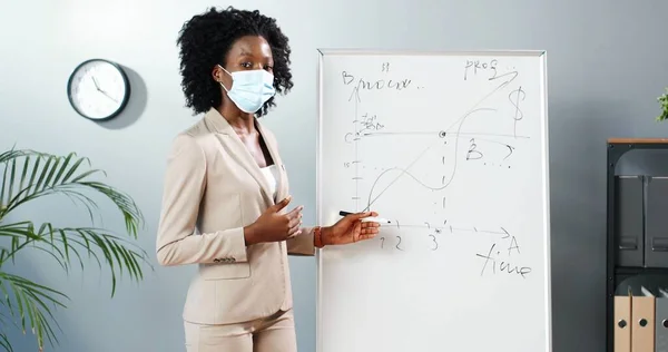 의료 마스크를 쓰고 있는 젊은 아프리카계 미국인 여성이 수업중에 칠판에 서서 물리나 기하학 법칙을 수업 시간에 알려 주고 있습니다. 세계적 인 컨셉이야. 코로나 바이러스가 있는 학교. 교육적 인 교육. — 스톡 사진