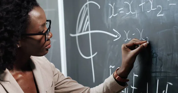 Primer plano de la joven profesora afroamericana en la escuela escribiendo fórmulas y leyes de mahemática en pizarra. Concepto escolar. Profesora de gafas explicando las leyes de física. — Foto de Stock