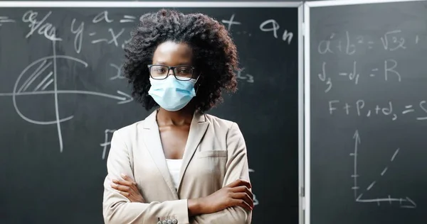 Porträt einer jungen afroamerikanischen Lehrerin mit Brille und medizinischer Maske, die in die Kamera blickt und sich im Klassenzimmer die Hände kreuzt. Tafel mit mathematischen Formeln im Hintergrund. Pandemische Schulbildung. — Stockfoto