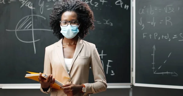 Retrato de una joven profesora afroamericana en gafas y máscara médica mirando a la cámara en clase y sosteniendo cuadernos. Pizarra con fórmulas sobre fondo. Educación pandémica. —  Fotos de Stock