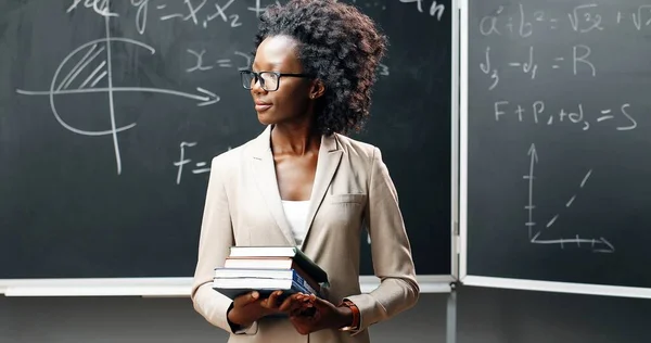 Portret van de jonge Afro-Amerikaanse lerares met een bril die in de klas naar de camera kijkt en schoolboeken vasthoudt. Schoolbord met formules op achtergrond. Schoolconcept. Boeken in handen van een vrouw. — Stockfoto