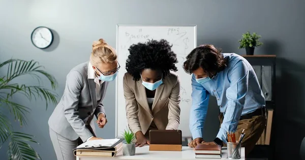 Three mixed-races male and females in medical mask standing in office and watching something on smartphone. Multiethnic teachers discussing online concept of studying via mobile phone. Man and women. — Stock Photo, Image