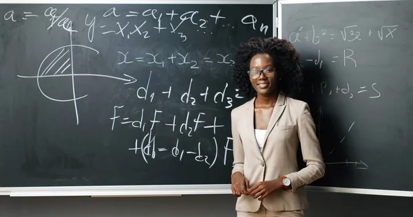 Portret van de jonge Afro-Amerikaanse lerares met een bril aan boord, schrijvend met krijt wiskunde of natuurkunde wetten en formules en glimlachend naar de camera in de klas. Onderwijsconcept. — Stockfoto
