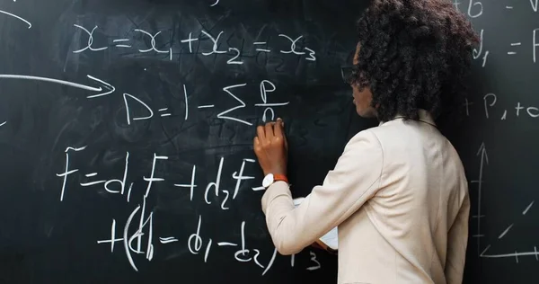 Terug blik op Afro-Amerikaanse jonge vrouw leraar op school schrijven formules en wiskunde wetten op schoolbord. Schoolconcept. Vrouwelijke docent in glazen die natuurkunde wetten uitlegt. Achterzijde. — Stockfoto