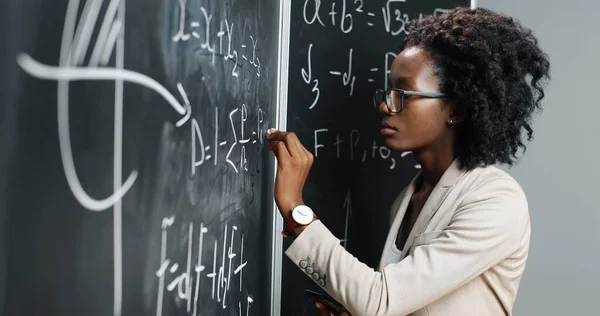 Jovem professora afro-americana na escola escrevendo fórmulas e leis matemáticas em quadro-negro. Conceito escolar. Professora de matemática feminina em óculos que explicam leis de física. Educação. — Fotografia de Stock