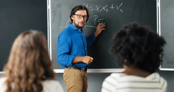 Caucasian man teacher at school writing formulas and mathematics laws on blackboard. School concept. Male lecturer in glasses explaining math laws to pupils. Educational concept.