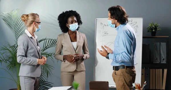 Caucasian African American females and male in medical masks talking and brainstorming. Pandemic corona concept. Mixed-races businessman and businesswomen in office. Multi ethnic man and women talk.