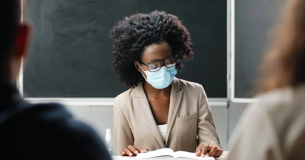 Joven profesora afroamericana en gafas y máscara médica sentada en la mesa de la escuela en clase, leyendo libros de texto y enseñando. Lección de literatura. Mujer educadora frente a estudiantes o alumnos. —  Fotos de Stock